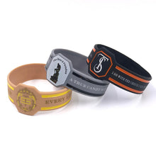 Three silicone wristbands with custom printed designs, displayed on a clean white background. One wristband is tan with yellow accents and a gold emblem; another is dark gray with silver accents and a black emblem; the third is black with orange accents and a white emblem.