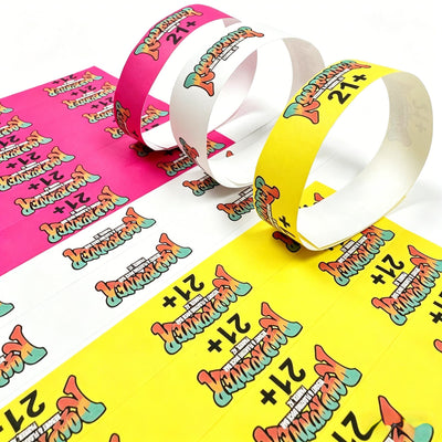 Three rolled Tyvek Wristbands in pink, white, and yellow, each printed with a colorful retro-style graphic and the text '21+' and 'The Prom Queen'.