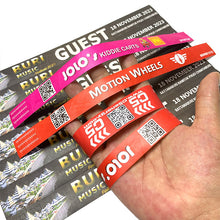 A hand holding four colorful event Tyvek Wristbands in front of printed event tickets. The wristbands are bright pink, orange-red, and orange, with white text and QR codes.