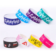 Six customizable Tyvek Wristbands in various colors (purple, blue, teal, black, white, yellow, pink) arranged on a plain white background. Each wristband features different printed designs, including text like 'Catering', 'TOP100 DTF', and '#Time to.