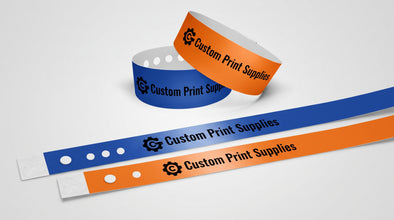 Two Custom Vinyl Wristbands, one blue and one orange, displayed on a light gray background with a second rolled orange wristband behind them. Each wristband features the text 'Custom Print Supplies' with a gear icon.